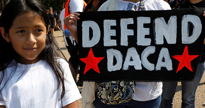 Young girl standing next to protest sign that reads, "Defend DACA"