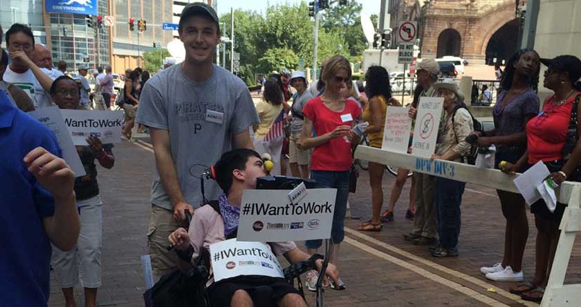People marching in Labor Day parade downtown. Young man is pushing another young man in a wheel chair, who is holding a sign that says, "I want to work."