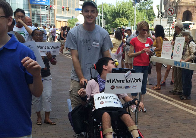 Street view during Labor Day Parade in Pittsburgh showing young man pushing another in a wheelchair who is holding a sign that says, #IWantToWork.