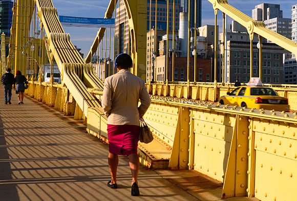 People walking on the sidewalk of a yellow bridge as they head into Pittsburgh.
