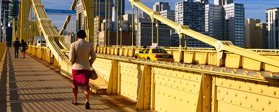 People walking on the sidewalk of a yellow bridge as they head into Pittsburgh.