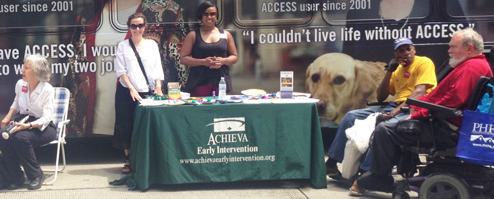Photo taken during the 3 Rivers Arts Festival celebrating the 25th anniversary of the Americans with Disabilities Act. Image shows several people in wheel chairs around an informational table provided by Achieva.