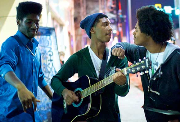 Three young African America men are singing on a city street. One is playing the guitar.