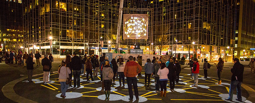 Nighttime image of a group of people interacting with the “Congregation” public art project in Market Square in Downtown Pittsburgh. This work was created by KMA and presented by Market Square Public Art.