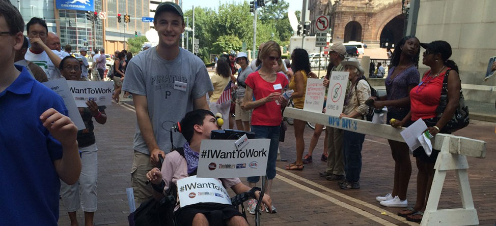 Street view during Labor Day Parade in Pittsburgh showing young man pushing another in a wheelchair who is holding a sign that says, "#IWantToWork.