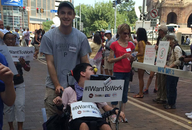 Street view during Labor Day Parade in Pittsburgh showing young man pushing another in a wheelchair who is holding a sign that says, "#IWantToWork.