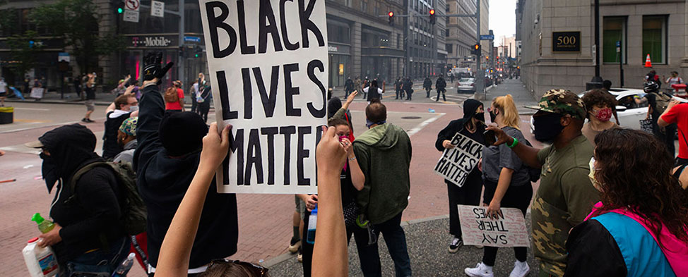 Both black and white people protesting Downtown on the death of George Floyd. Everyone is wearing masks because of the pandemic and people are carrying "Black Lives Matter" signs. The police are in the background.