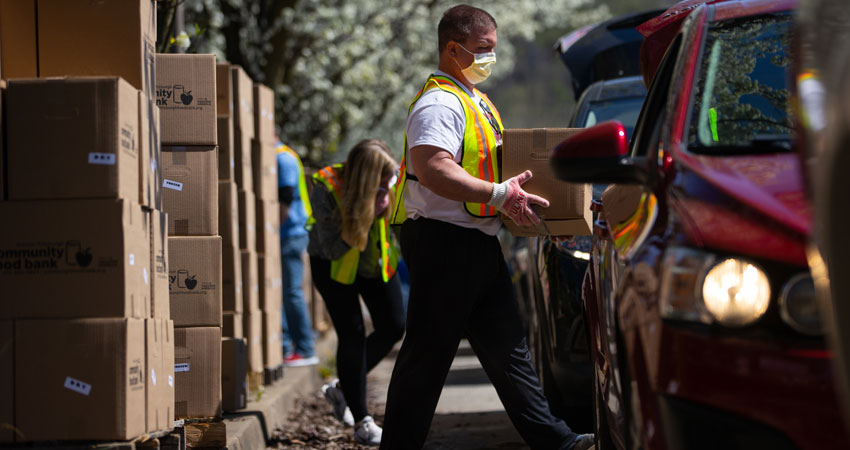 Two volunteers wearing face masks and gloves are loading boxes of food from the Foodbank into the cars of people lined up to receive them.