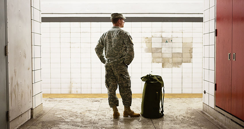 soldier standing in a hallway with his duffle bag next to him on the floor staring at a blank tile wall