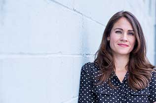 Betty Cruz, with long brown hair, leaning against a white concrete block wall.