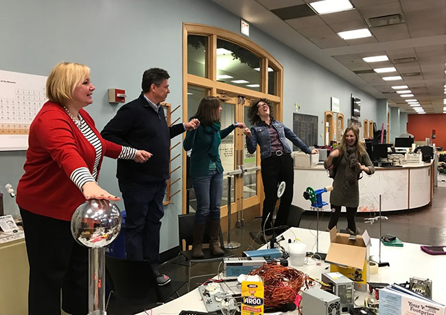 Five cohort teachers are playing with a vandagraph generator during their visit to YouMedia at the Chicago Public Library. They are laughing and holding hands.